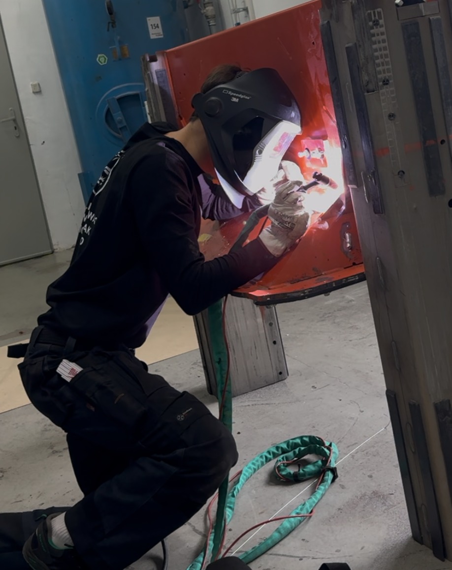 Pracownik w masce spawalniczej, w trakcie spawania elementu konstrukcyjnego. Widoczne iskry i narzędzia. Ujęcie z boku, z detalami wyposażenia ochronnego.