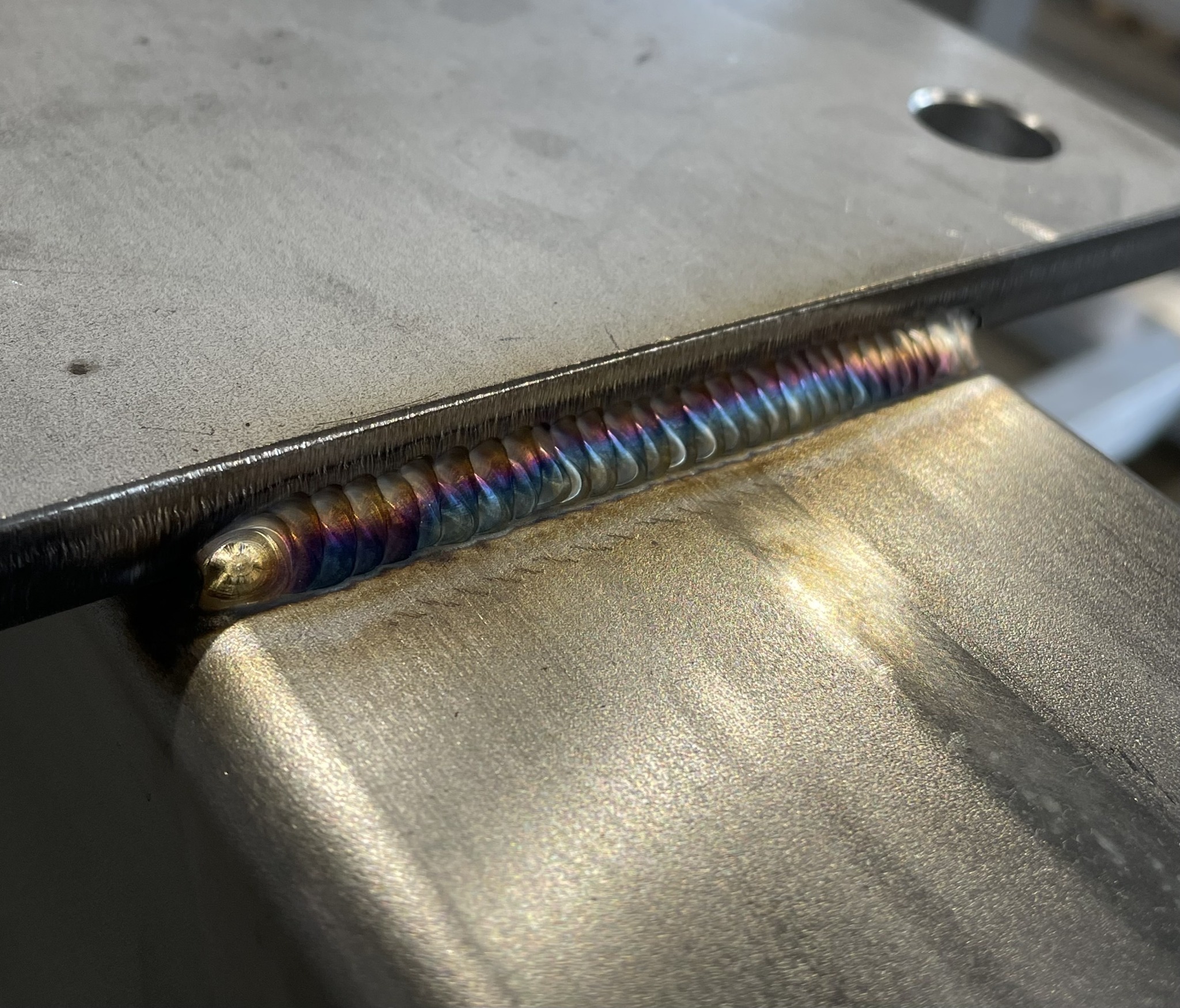 Precyzyjne, tęczowe spawy na metalowej konstrukcji. Widoczne regularne, drobne fale spoiny. Powierzchnia metalu delikatnie szlifowana, z widocznymi śladami obróbki.