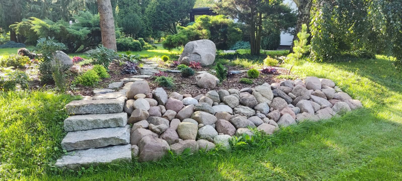 Ogród z kamiennymi schodami i rabatą z kamieni. W tle drzewa i krzewy. Ujęcie w słoneczny dzień, z perspektywy trawnika. Elementy małej architektury ogrodowej.