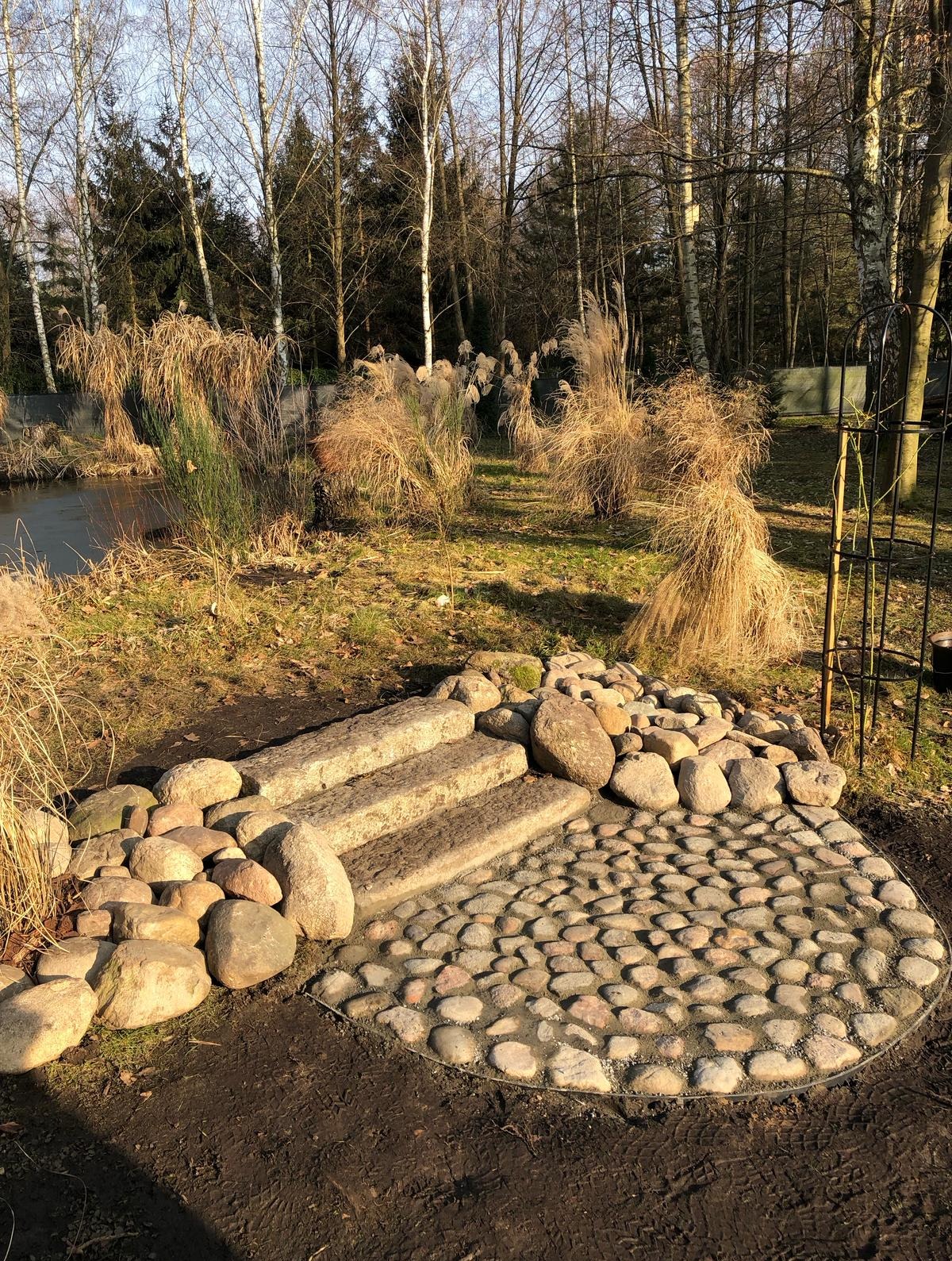 Kamienne schody i okrągła ścieżka z bruku w ogrodzie, otoczone ozdobnymi trawami i drzewami. Elementy małej architektury ogrodowej w naturalnym otoczeniu.