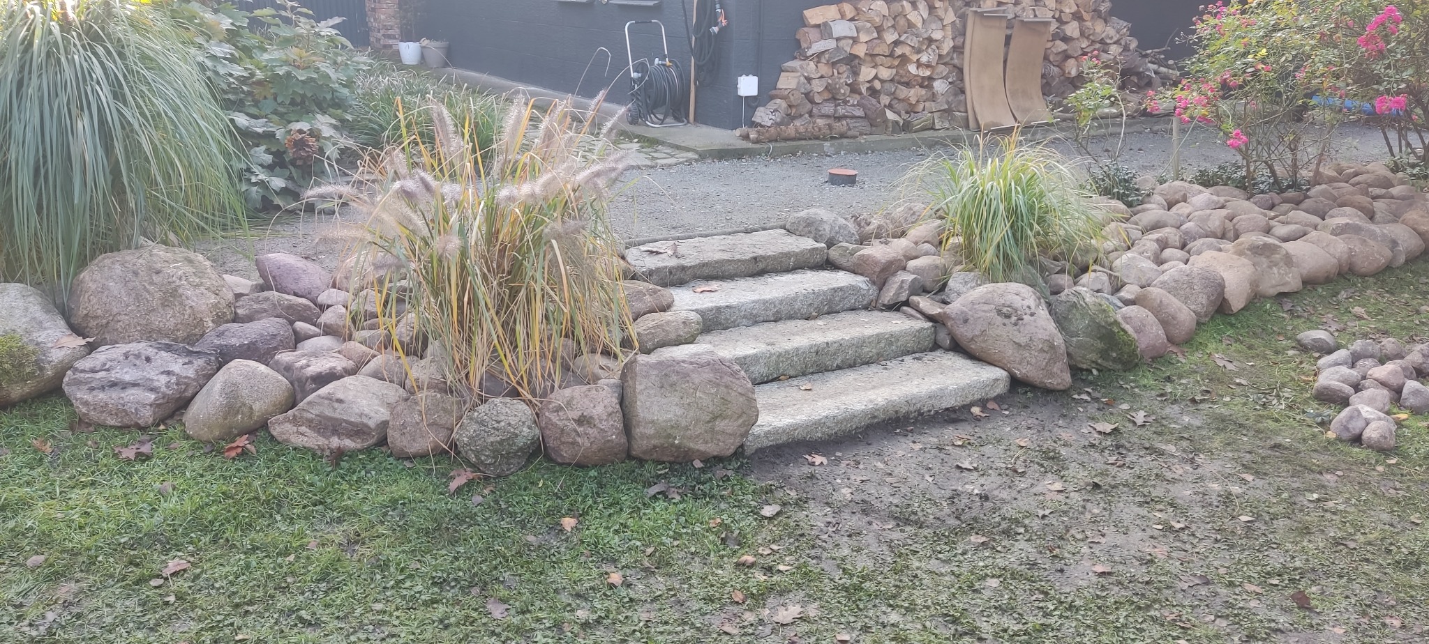 Kamienne schody ogrodowe z ozdobnymi głazami i trawami, prowadzące przez zielony trawnik. W tle widoczne drewno i roślinność.