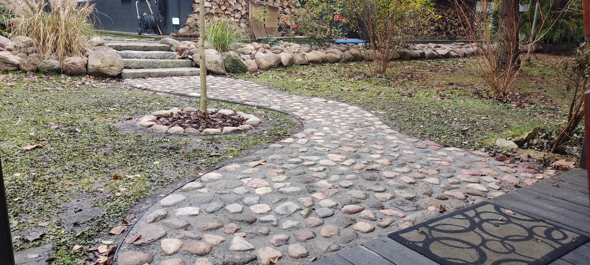 Kręta ścieżka z kamienia polnego prowadzi przez ogród, otaczając małe drzewko. Kamienne schody i murki z kamieni w tle. Dywanik przed wejściem na drewniany taras.