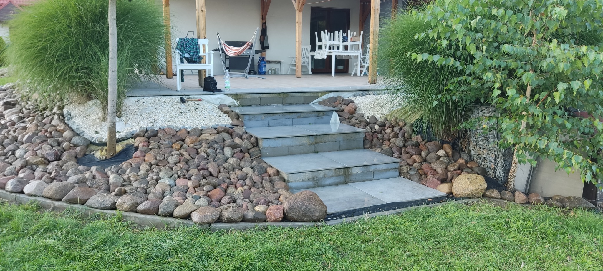 Kamienne schody prowadzące na taras w Obornikach Śląskich. Dekoracyjne kamienie i roślinność po bokach. Widoczny fragment domu z hamakiem i meblami.