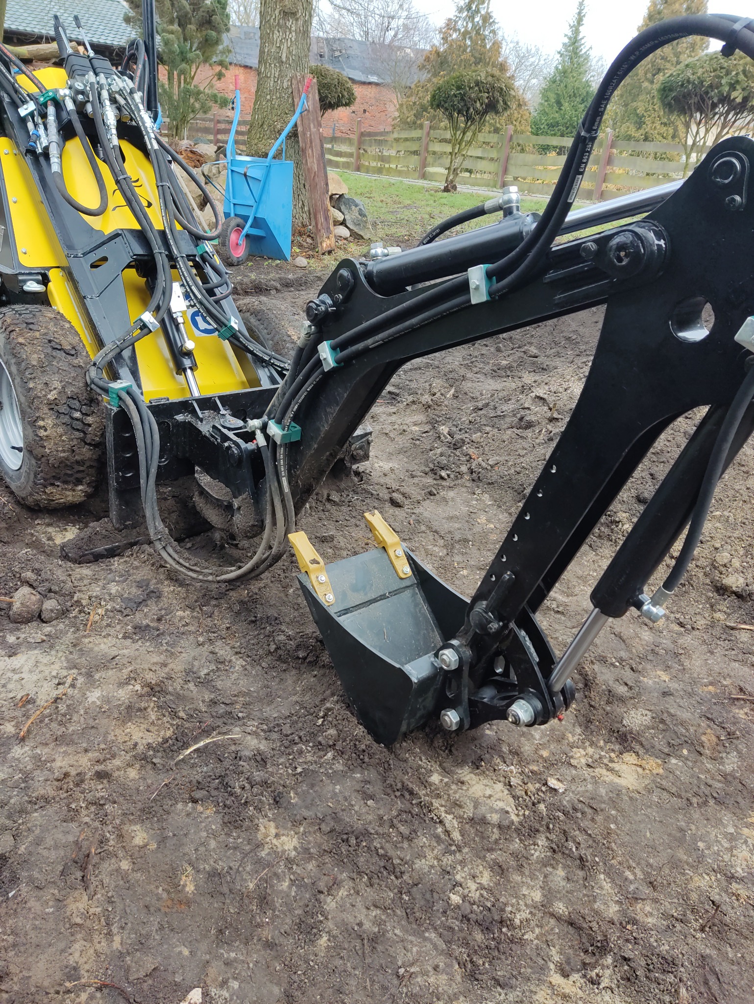 Żółto-czarna minikoparka z łyżką w ziemi, widok z bliska. W tle widać drzewa i budynek. Scena sugeruje przygotowanie terenu pod prace ziemne.