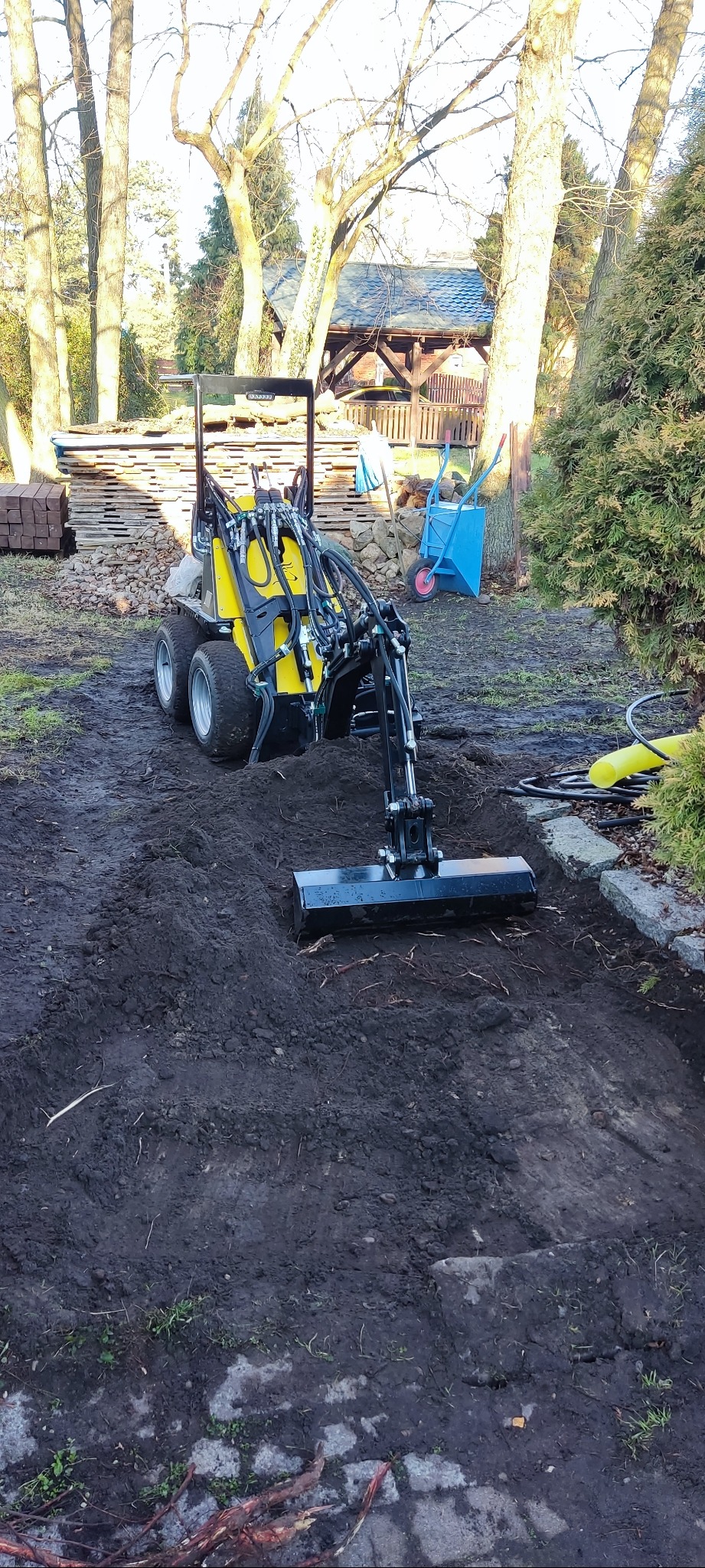 Żółta miniładowarka na grząskim terenie, przygotowująca podłoże pod prace ogrodowe. W tle palety z kostką, taczka i altana ogrodowa.