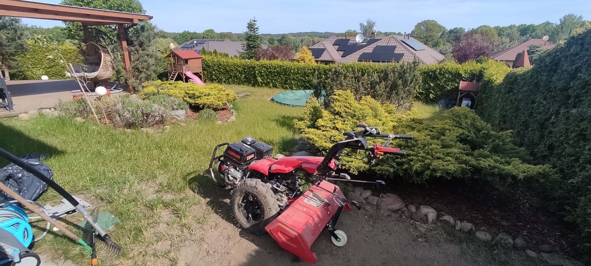 Czerwony glebogryzarek spalinowy na tle bujnego ogrodu z domkiem dla dzieci, huśtawką i zadbanym trawnikiem. W tle dom z panelami słonecznymi.