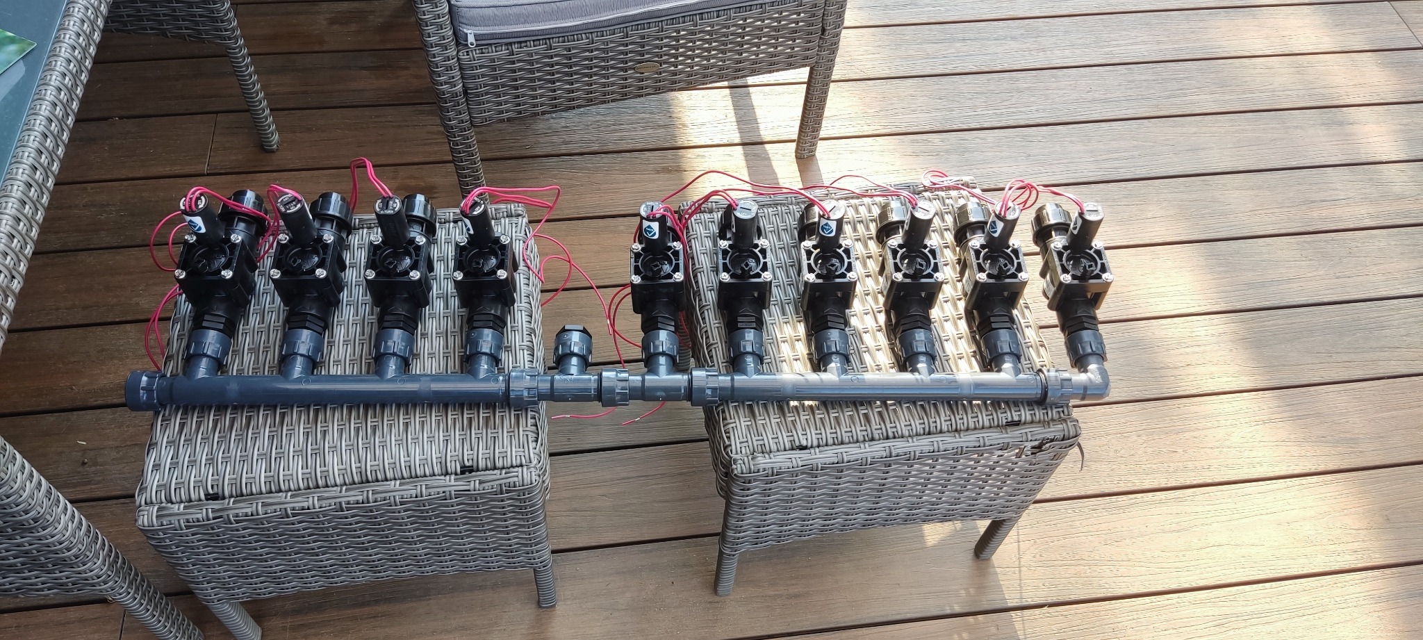 Zmontowany system nawadniania z czarnymi zraszaczami i szarymi rurami PVC, ułożony na dwóch plecionych stołkach na drewnianym tarasie, gotowy do instalacji.