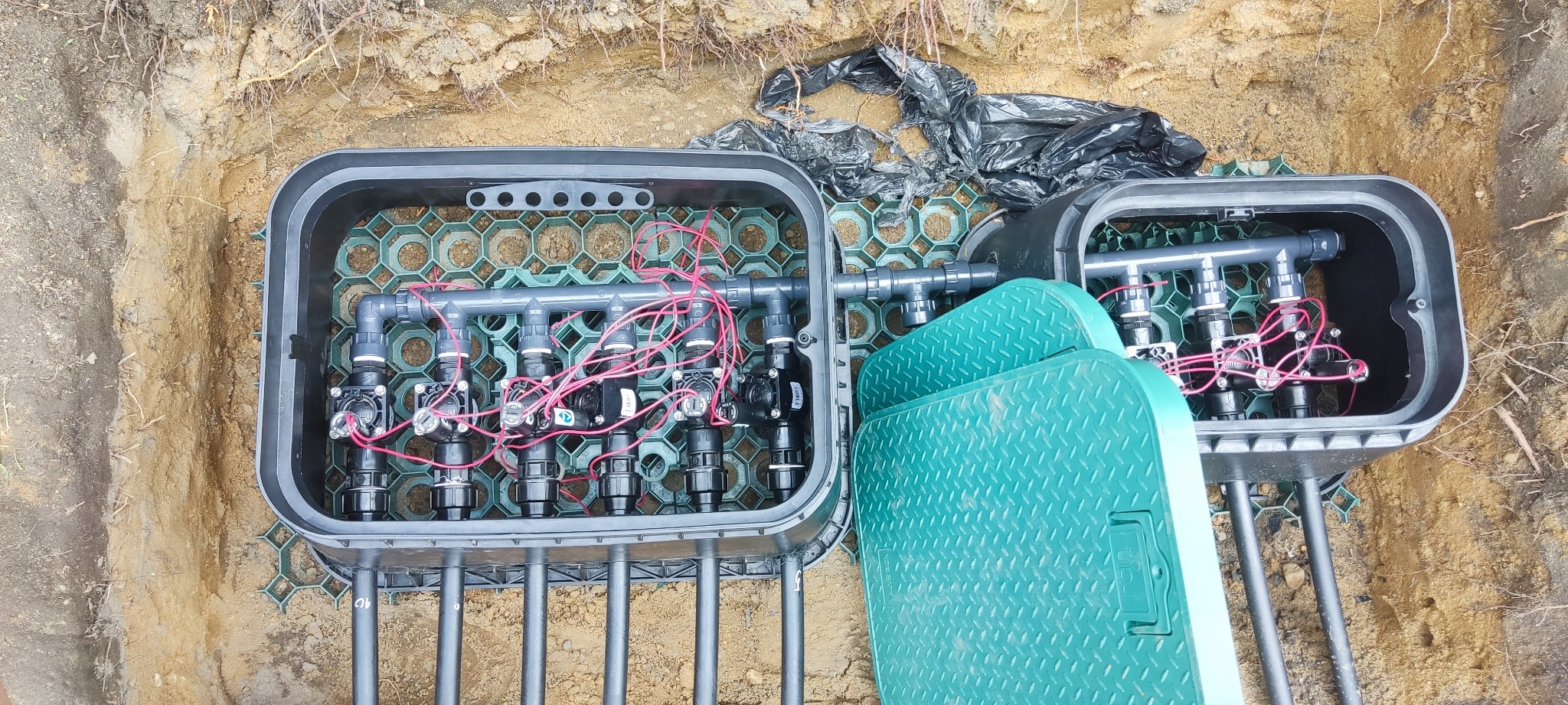 Ziemny wykop z zainstalowanym systemem nawadniania w szarej skrzynce z zielonymi pokrywami. Widoczne rury, zawory i okablowanie. Czarna folia w tle.