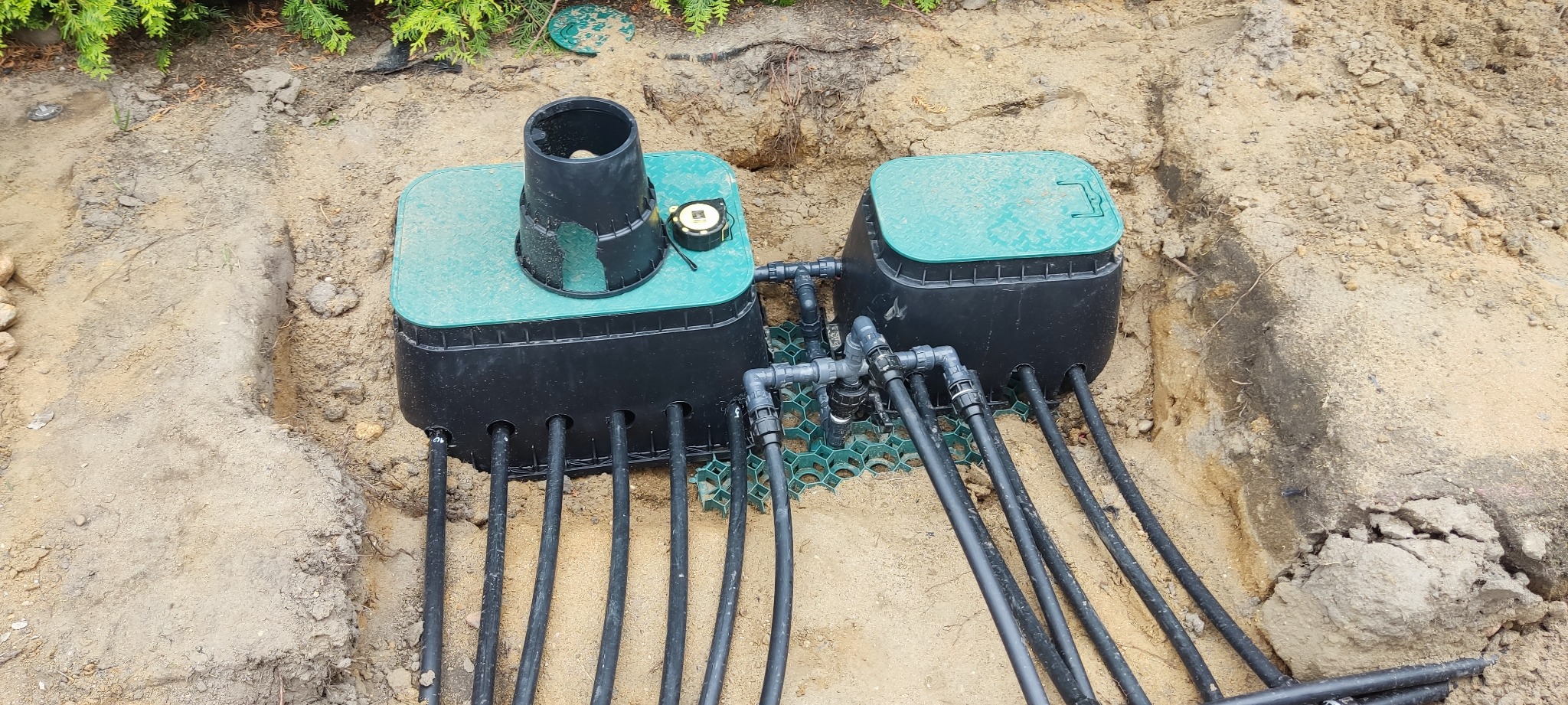 Podziemny system nawadniania w trakcie instalacji: zielone skrzynki z czarnymi rurami na tle ziemi. Widoczna taśma miernicza na jednej ze skrzynek.