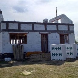 Grzegorz Mołas - Budowa domu jednorodzinnego w stanie surowym otwartym: białe bloczki H+H, szare nadproża, drabina oparta o ścianę, widoczne drzewa w tle.