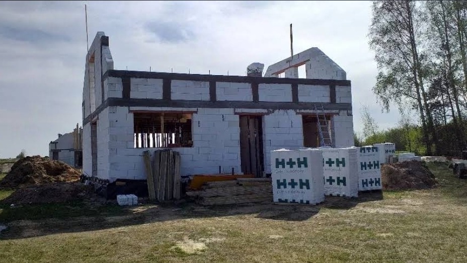 Budowa domu jednorodzinnego w stanie surowym otwartym: białe bloczki H+H, szare nadproża, drabina oparta o ścianę, widoczne drzewa w tle.