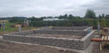 Wzniesione mury fundamentowe z szarej bloczków betonowych na tle zieleni i pochmurnego nieba. Widoczna instalacja kanalizacyjna. Częściowo ułożone bloczki.