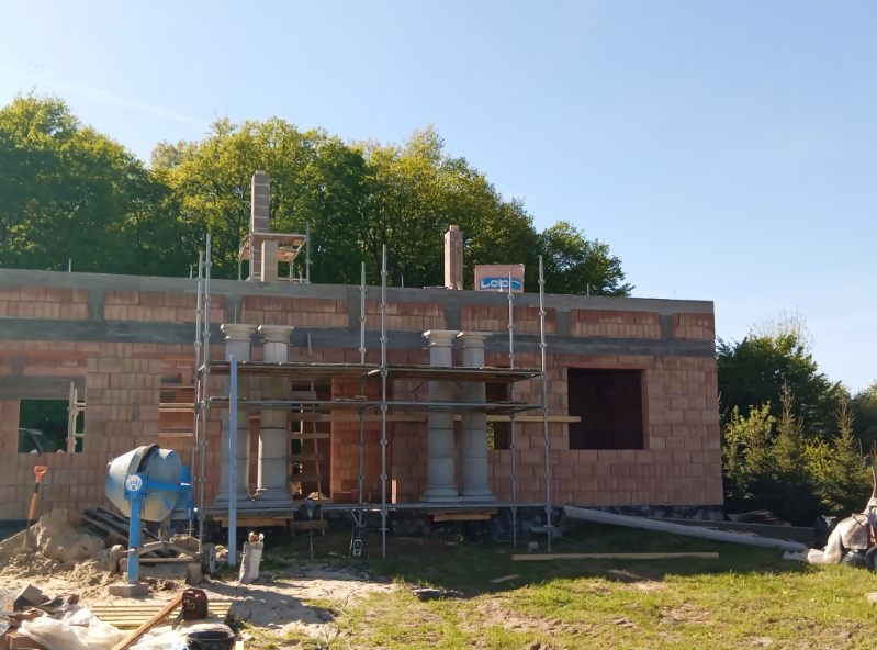 Wznoszenie murów domu z cegły, widoczne rusztowania i elementy dekoracyjne w stylu kolumn. Na pierwszym planie betoniarka, w tle drzewa.