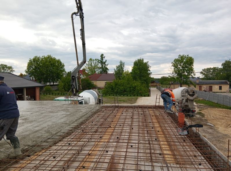 Zalewanie fundamentów domu: beton z betoniarki wylewany na zbrojenie, widoczni pracownicy w odzieży roboczej i pompa do betonu. Na drugim planie zabudowa.