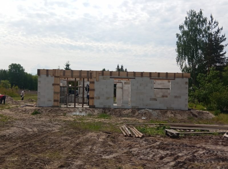 Wznoszenie ścian z betonu komórkowego domu jednorodzinnego. Widoczne drewniane nadproża okienne i drzwiowe. Na pierwszym planie sterta drewna budowlanego.