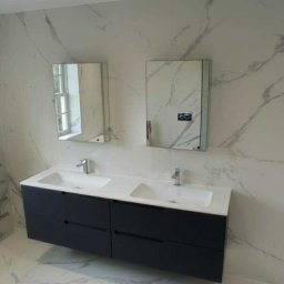 FI-WIT INTERIORS S.C. - Wykończona łazienka z marmurowymi płytkami, podwójną umywalką z szafką w kolorze granatowym, lustrami i grzejnikiem drabinkowym. Widok na zewnątrz przez okno.