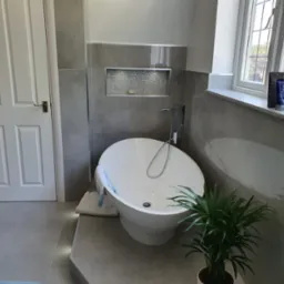 Nowoczesna łazienka z wolnostojącą wanną, szarą glazurą i wnęką z mozaiką. Minimalistyczny design podkreśla roślina doniczkowa i okno z widokiem na zieleń.