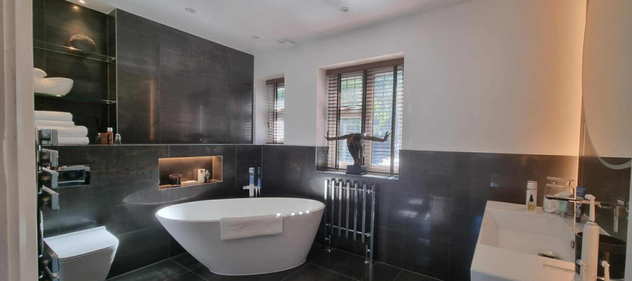 Nowoczesna łazienka z czarnymi płytkami, wolnostojącą wanną i drewnianymi żaluzjami. Dekoracyjna rzeźba przy oknie, minimalistyczne oświetlenie.