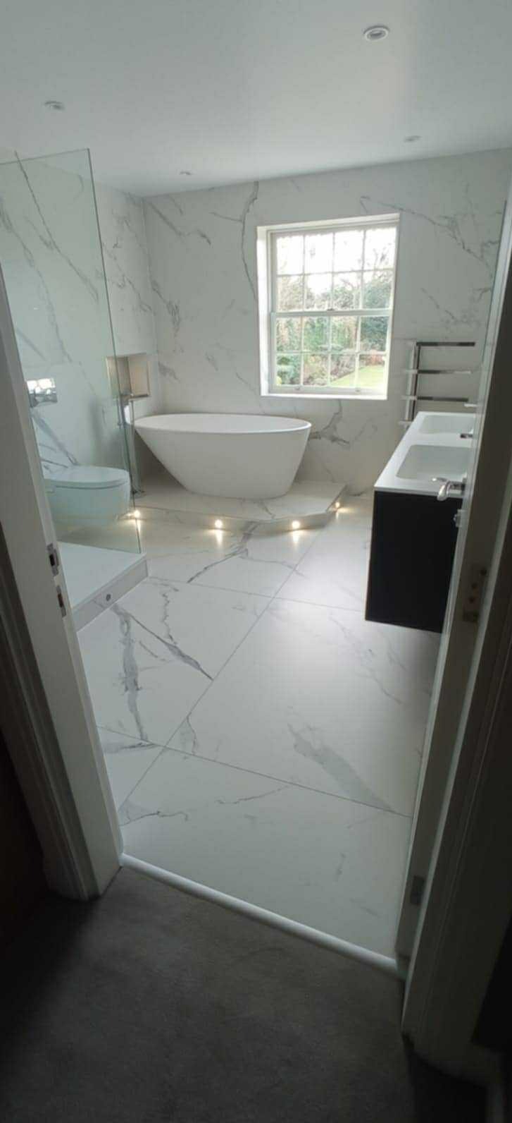 Elegancka łazienka w bieli z marmurowym wzorem na płytkach, owalna wanna, kabina prysznicowa ze szkła i podświetlenie ledowe pod wanną tworzą nowoczesny wystrój.