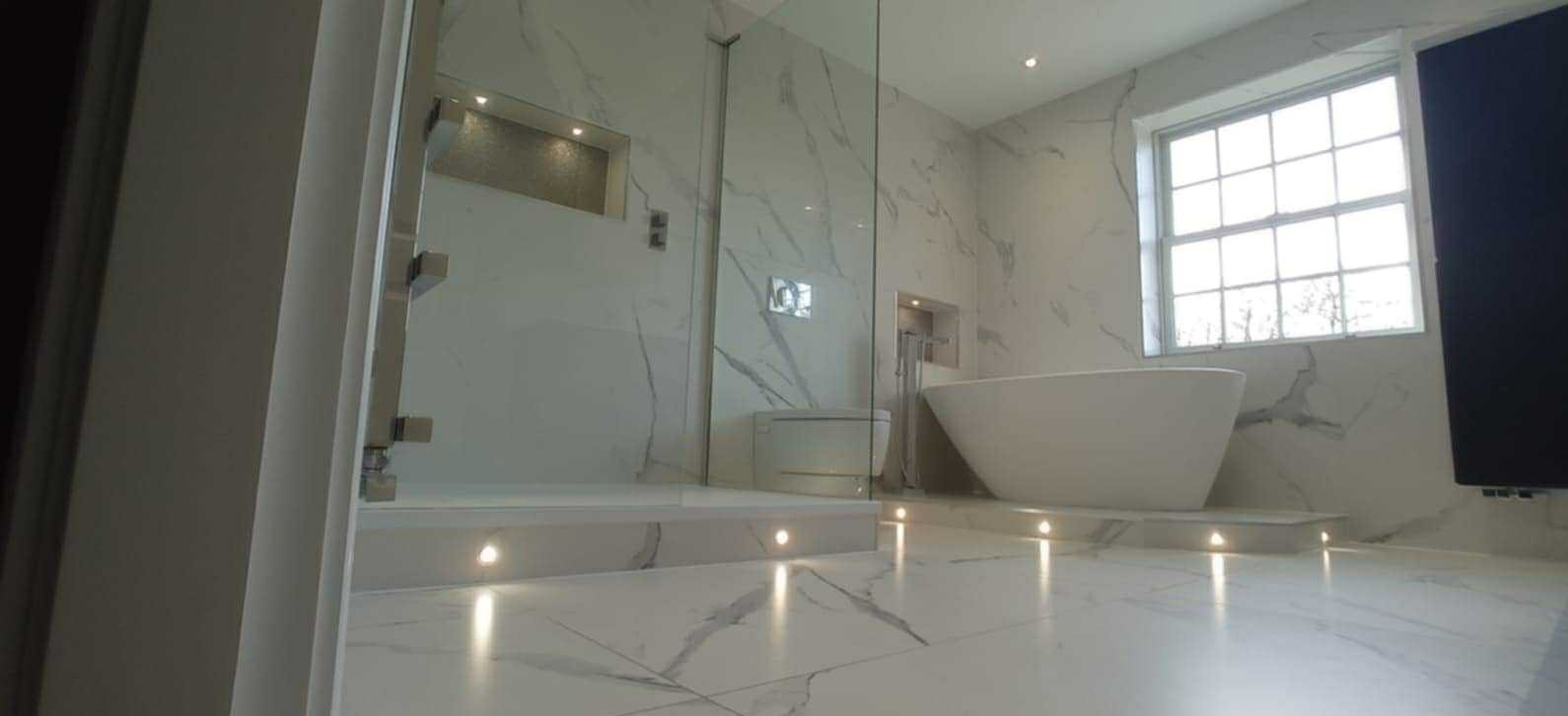 Elegancka łazienka z marmurowymi płytkami, przeszkloną kabiną prysznicową i wolnostojącą wanną pod oknem. Podświetlenie LED w podłodze. Nowoczesny design.