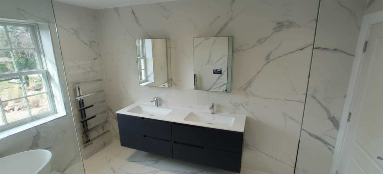 Wykończona łazienka z marmurowymi płytkami, podwójną umywalką z szafką w kolorze granatowym, lustrami i grzejnikiem drabinkowym. Widok na zewnątrz przez okno.