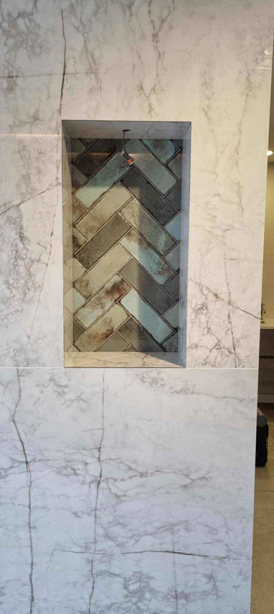 Wykończona wnęka ścienna w łazience z mozaiką ułożoną w jodełkę, otoczona płytami imitującymi marmur. Widoczny fragment ściany i podłogi.