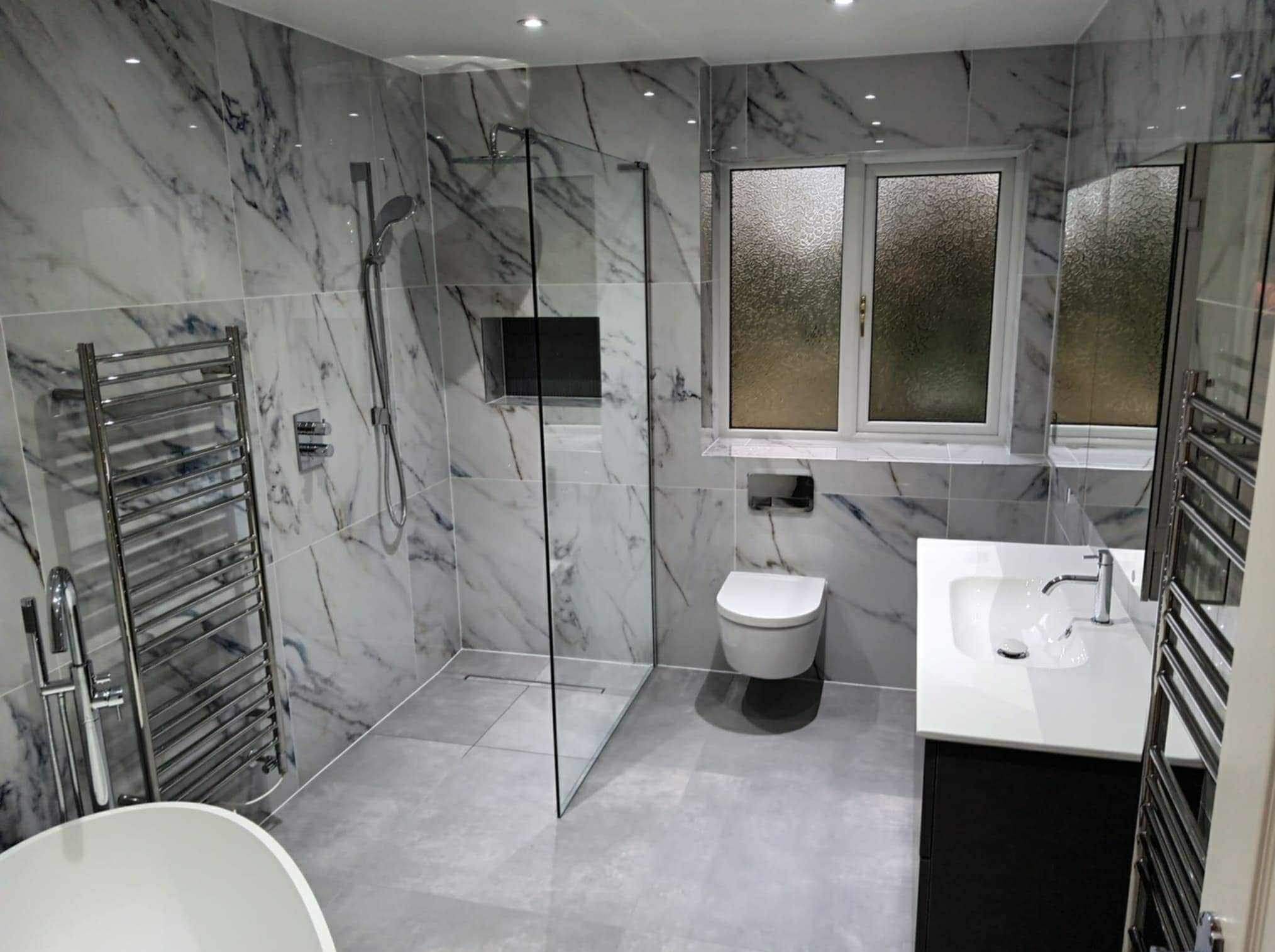 Elegancka łazienka po remoncie z marmurowymi płytkami, kabiną prysznicową typu walk-in, grzejnikiem drabinkowym i nowoczesną armaturą, oświetlona punktowo.