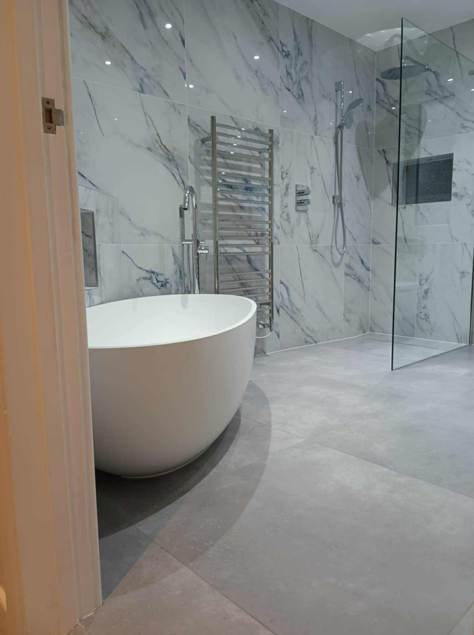 Elegancka łazienka po remoncie: wolnostojąca wanna, marmurowe płytki na ścianach, szklana kabina prysznicowa i grzejnik drabinkowy, widok zza uchylonych drzwi.