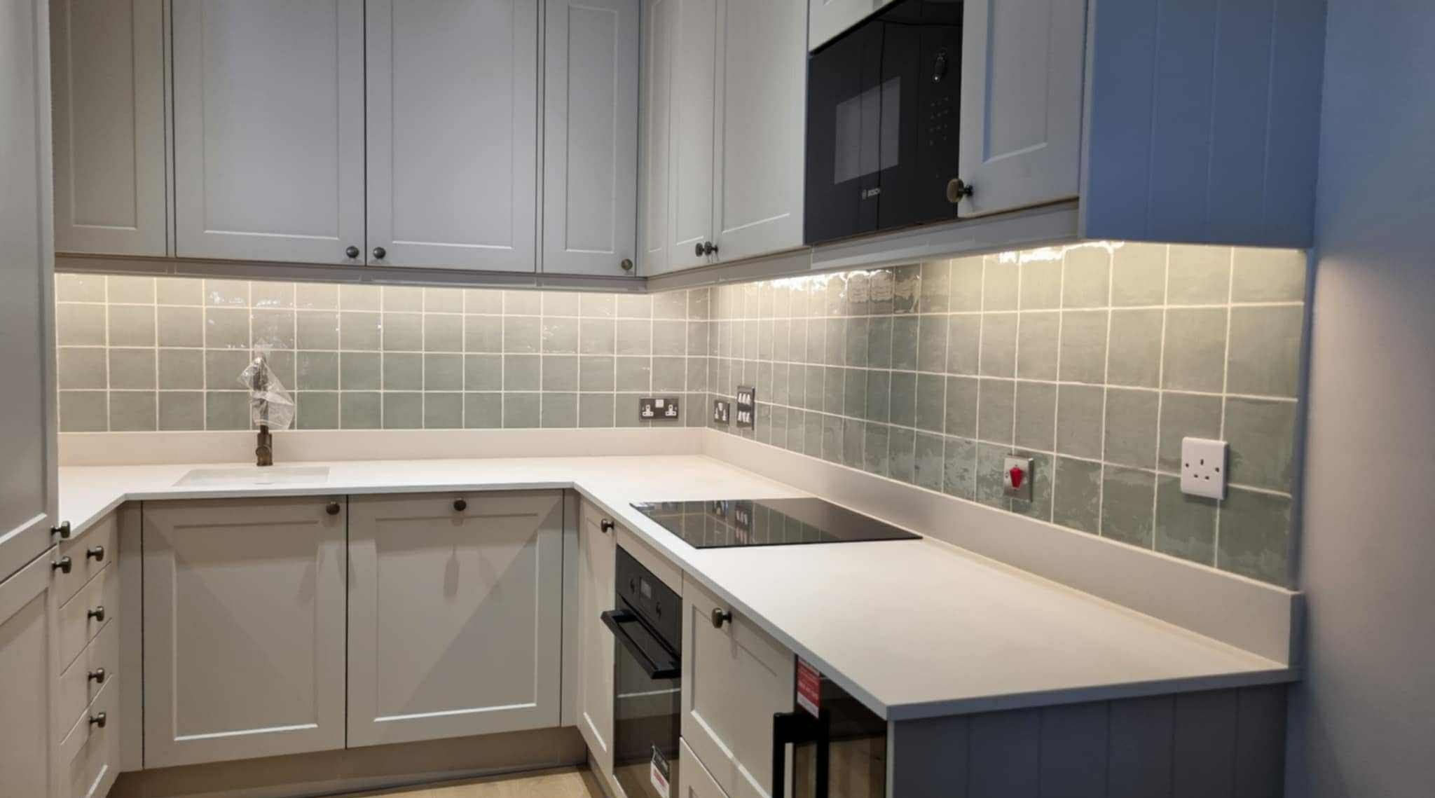 Jasna kuchnia w stylu angielskim z szarymi szafkami, płytkami w odcieniach zieleni i białym blatem. Widoczne gniazdka elektryczne i płyta indukcyjna.