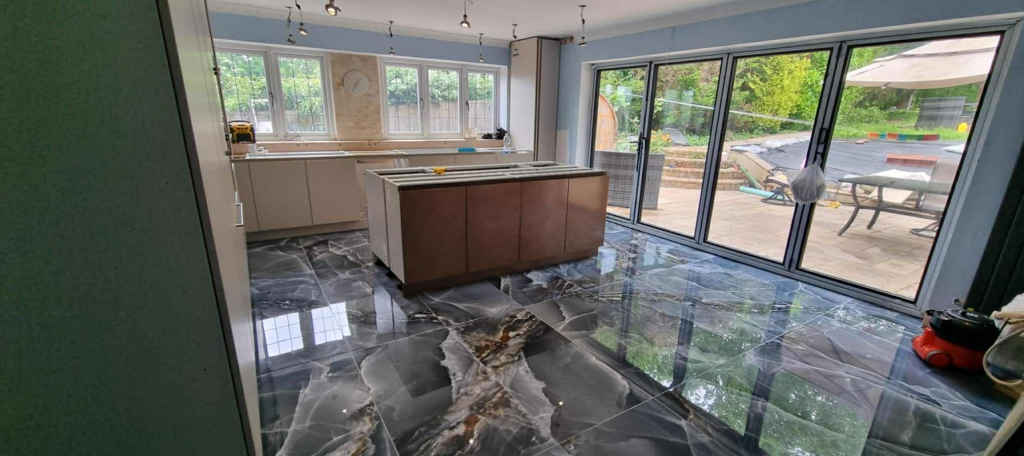 Wykończona kuchnia z marmurową podłogą i wyspą. Widoczne duże okna z widokiem na ogród. Częściowo widoczne szafki kuchenne i sprzęt. Oświetlenie sufitowe.