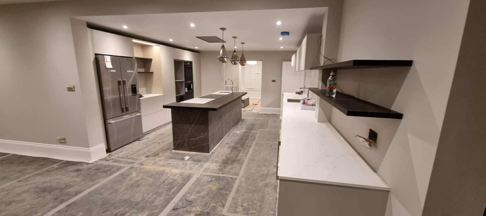 Nowoczesna, jasna kuchnia w trakcie wykańczania: marmurowa wyspa, zabudowa meblowa, płyty na podłodze z fugami. Widoczne elementy instalacji elektrycznej.