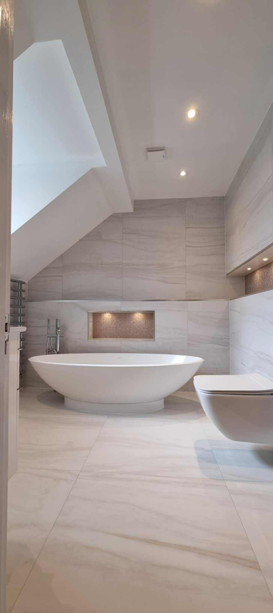 Elegancka łazienka w jasnych, marmurkowych płytkach. Wolnostojąca wanna i podwieszana toaleta. Wnęka z mozaiką i oświetleniem. Minimalistyczny design.