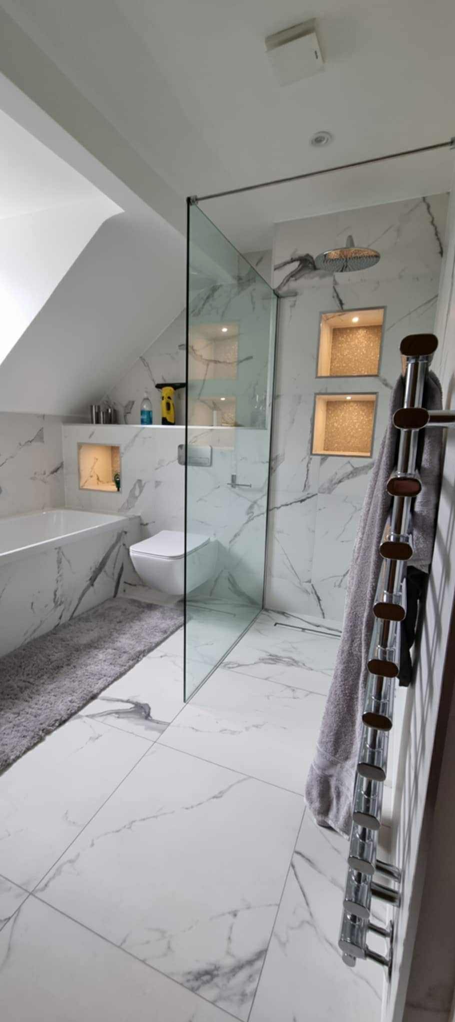 Elegancka łazienka na poddaszu z marmurowymi płytkami, szklaną kabiną prysznicową typu walk-in i designerskim grzejnikiem drabinkowym z ręcznikiem.