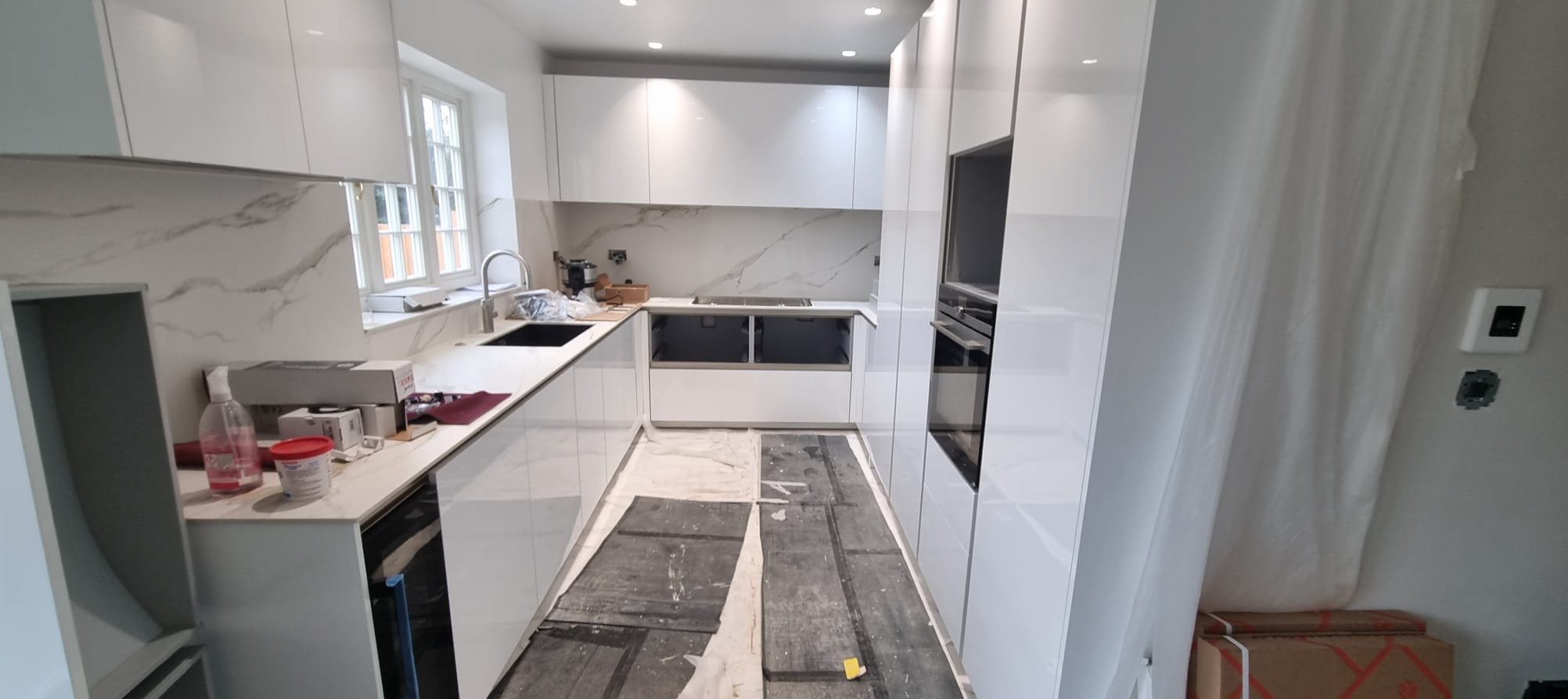 Nowoczesna, biała kuchnia z marmurowym blatem i backsplashiem. Widoczne elementy montażu, podłoga zabezpieczona folią. Minimalistyczny design, jasne oświetlenie.