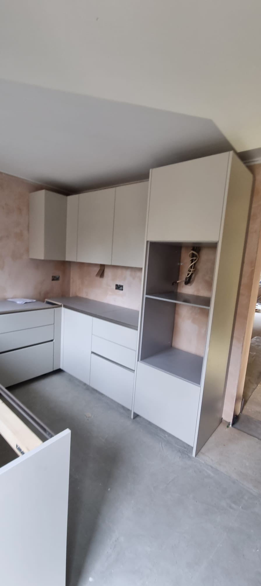 Jasna zabudowa kuchenna w trakcie montażu. Widoczne szafki wiszące i stojące, surowe ściany. Częściowo wysunięta szuflada. Stan surowy podłogi.