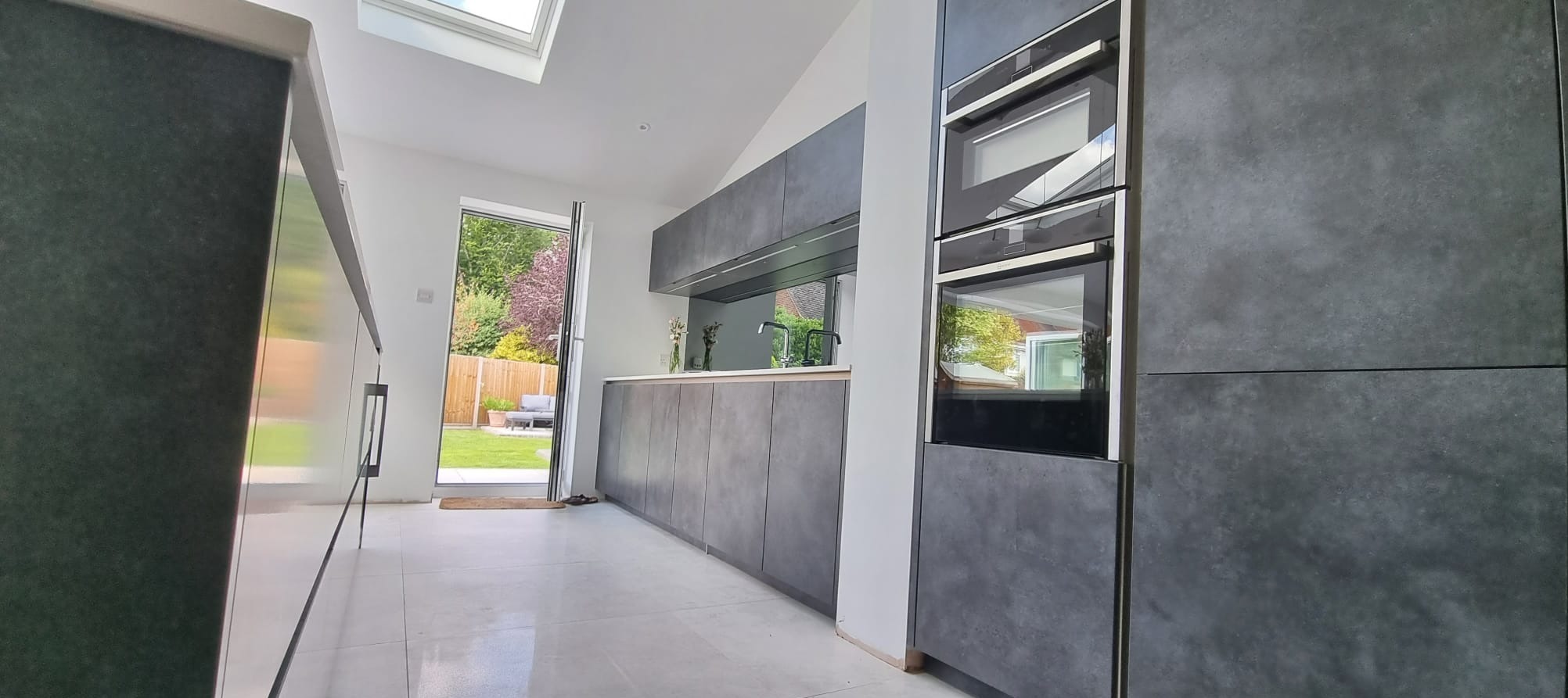 Nowoczesna kuchnia z szarymi matowymi frontami szafek, wbudowanymi piekarnikami i widokiem na ogród przez otwarte drzwi balkonowe.