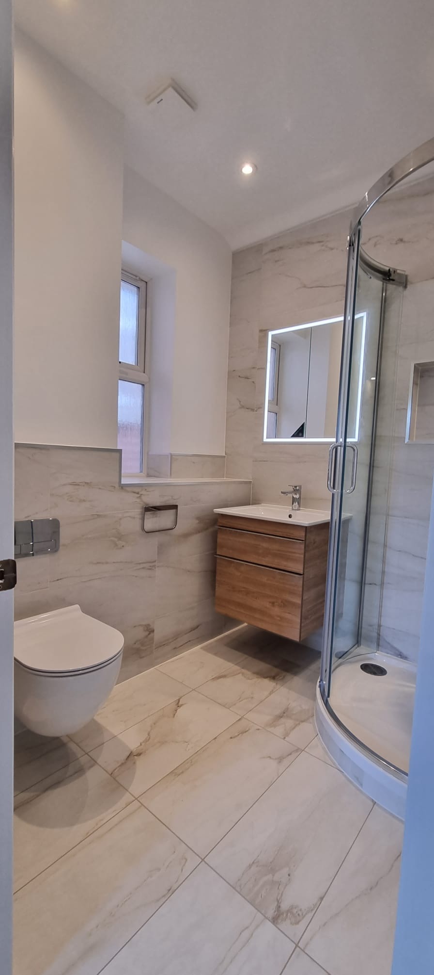 Wykończona łazienka z kabiną prysznicową, podwieszaną szafką z umywalką i podświetlanym lustrem. Biało-beżowe płytki imitujące marmur na podłodze i ścianach.