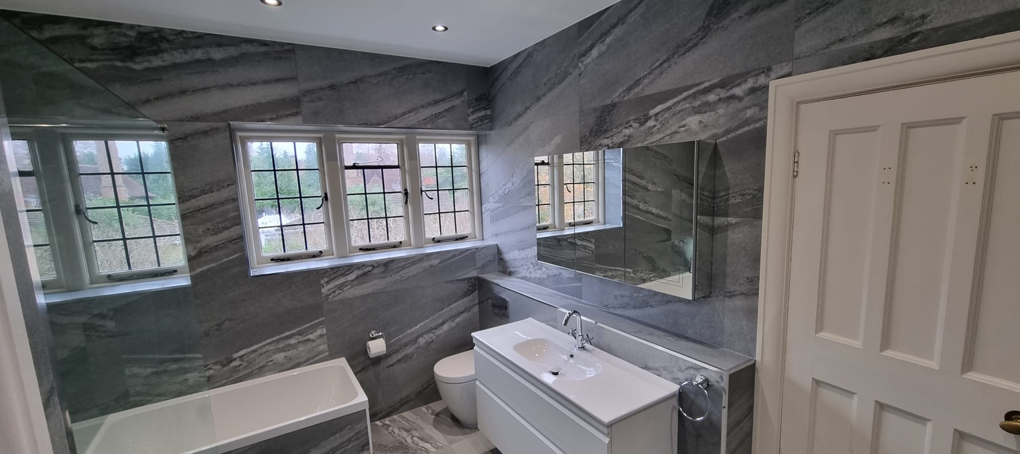 Elegancka łazienka z szarymi płytkami imitującymi marmur, biała armatura i okno z widokiem na zieleń. Lustro z szafką nad umywalką.