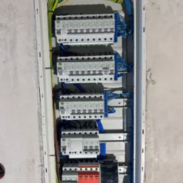 Rozdzielnica elektryczna z zabezpieczeniami, widoczne przewody w kolorach: niebieskim, zielono-żółtym, brązowym. Zamontowana w surowej ścianie.