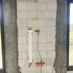 BT Power - Ściana z betonu komórkowego z widocznymi przewodami elektrycznymi w peszlach i puszkami instalacyjnymi. Widoczne okno i fragment instalacji podtynkowej.