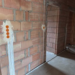 BT Power - Instalacja elektryczna w trakcie montażu w nowym budynku z czerwonej cegły. Widoczne kable w białych listwach i pomarańczowe puszki pod gniazdka.