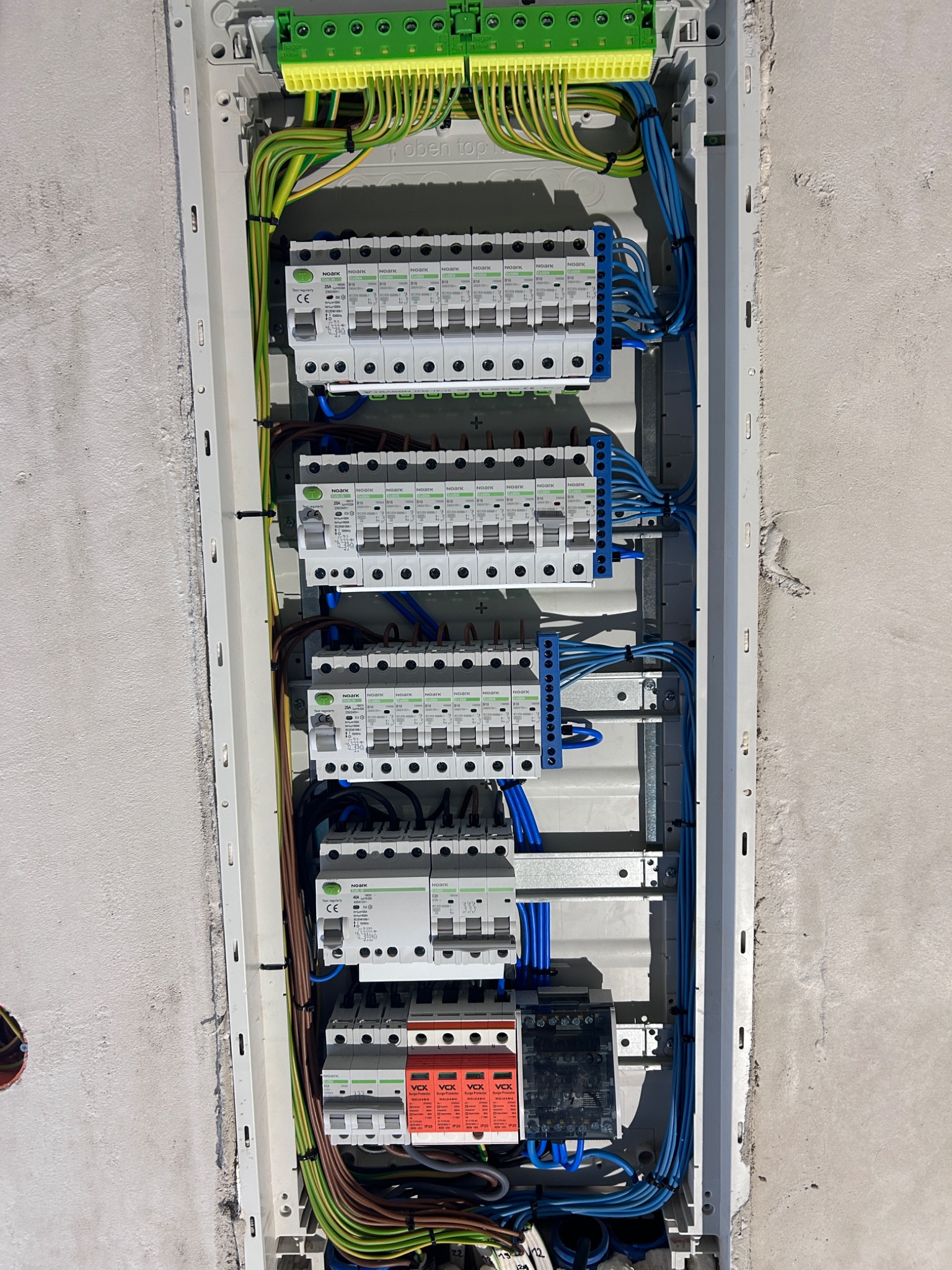 Rozdzielnica elektryczna z zabezpieczeniami, widoczne przewody w kolorach: niebieskim, zielono-żółtym, brązowym. Zamontowana w surowej ścianie.
