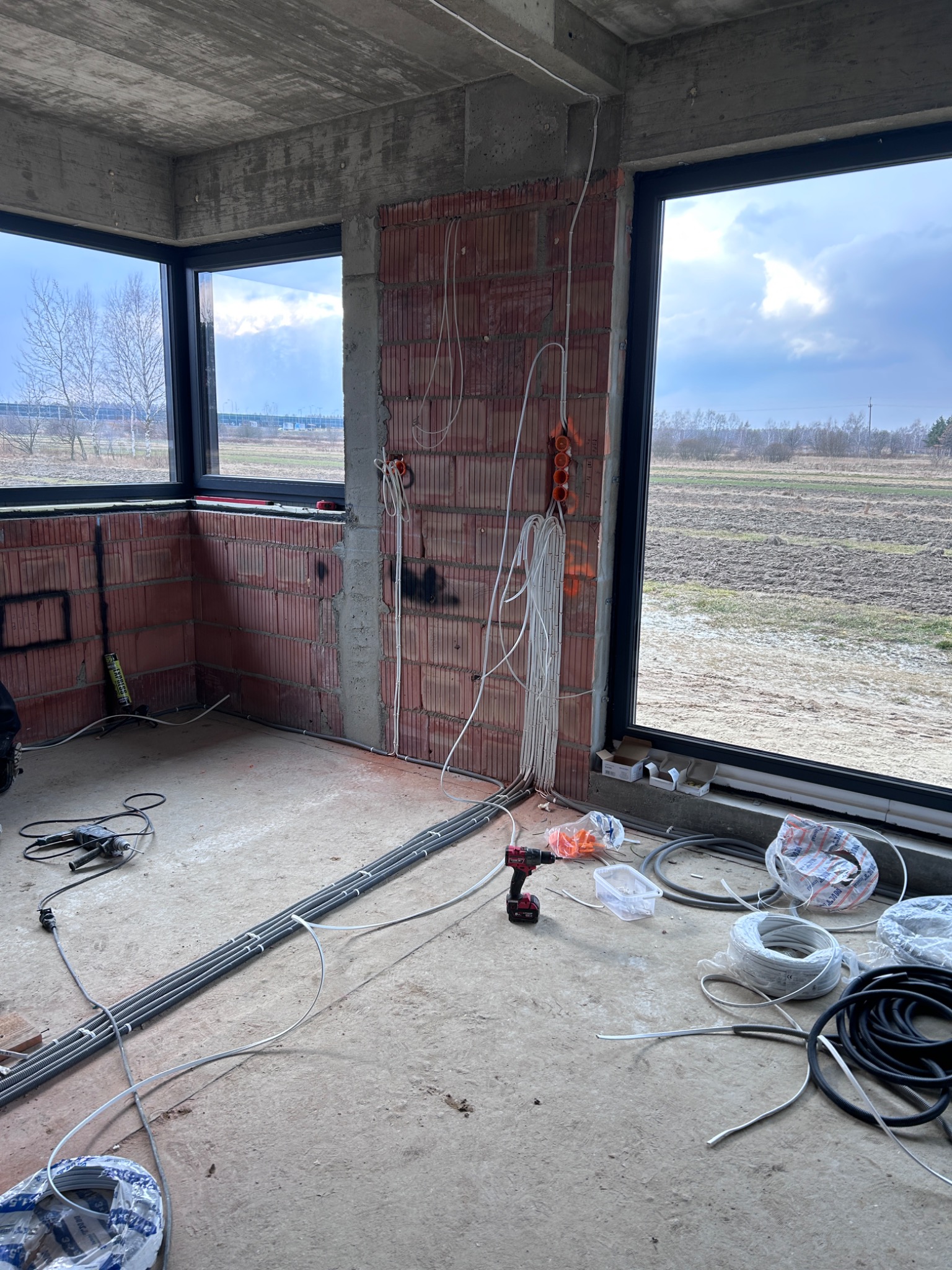 Wnętrze w stanie surowym z widocznymi przewodami elektrycznymi prowadzonymi po ścianach z cegły oraz ułożonymi na podłodze, narzędzia pracy i materiały budowlane.