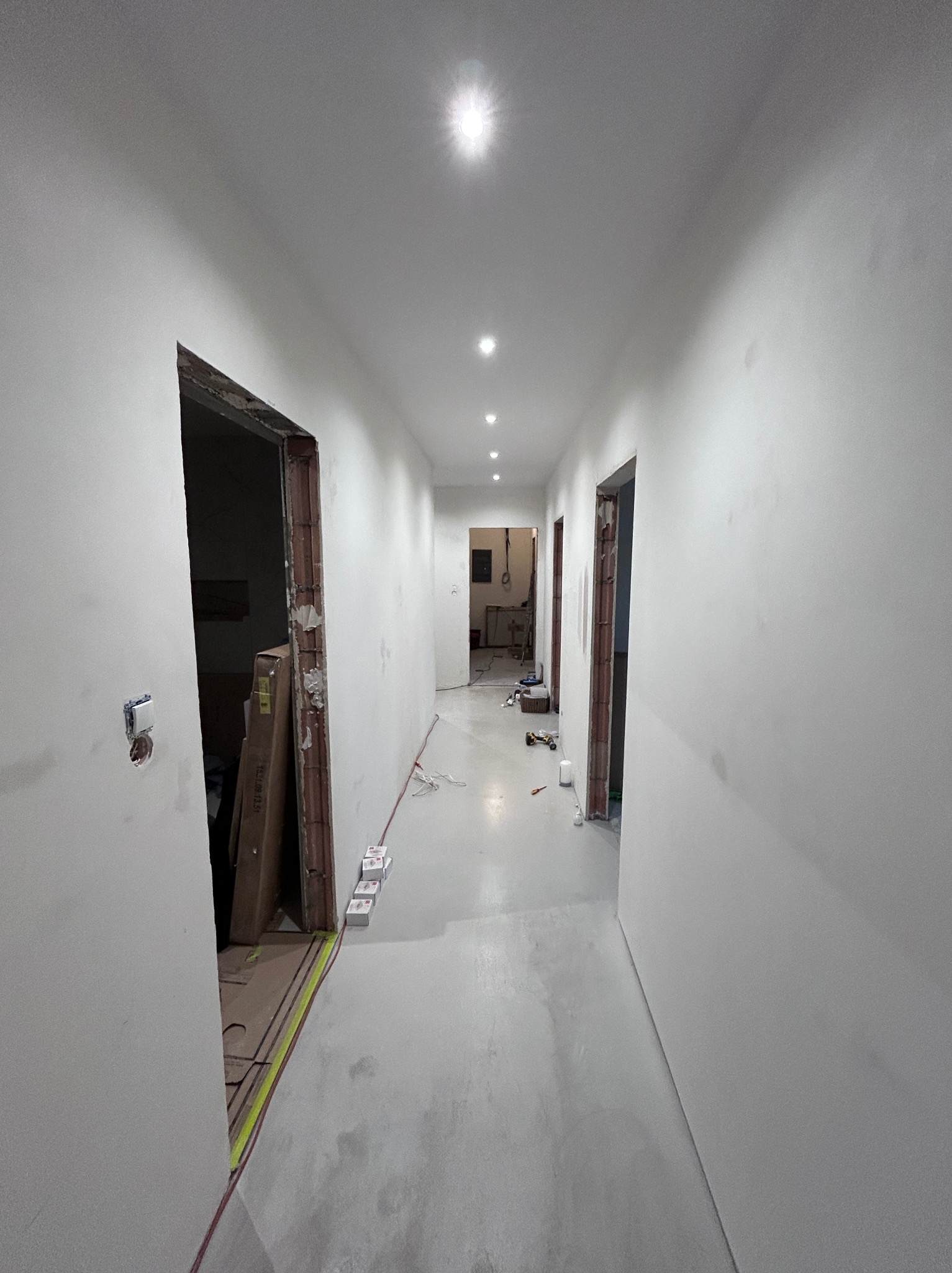 Korytarz w trakcie remontu: ściany po gładzi, widoczne puszki elektryczne, sufit z zamontowanym oświetleniem punktowym. Częściowo wykończone ościeżnice.