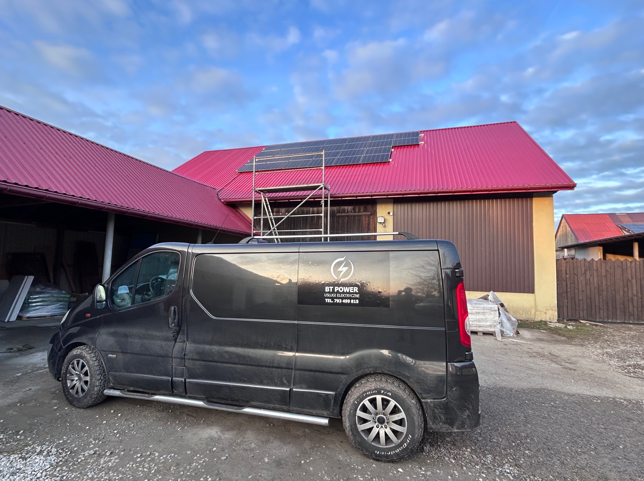 Czarny van z logo firmy elektrycznej na tle budynku z panelami słonecznymi na dachu i rusztowaniem. Widoczne materiały budowlane. Dzień, pochmurne niebo.