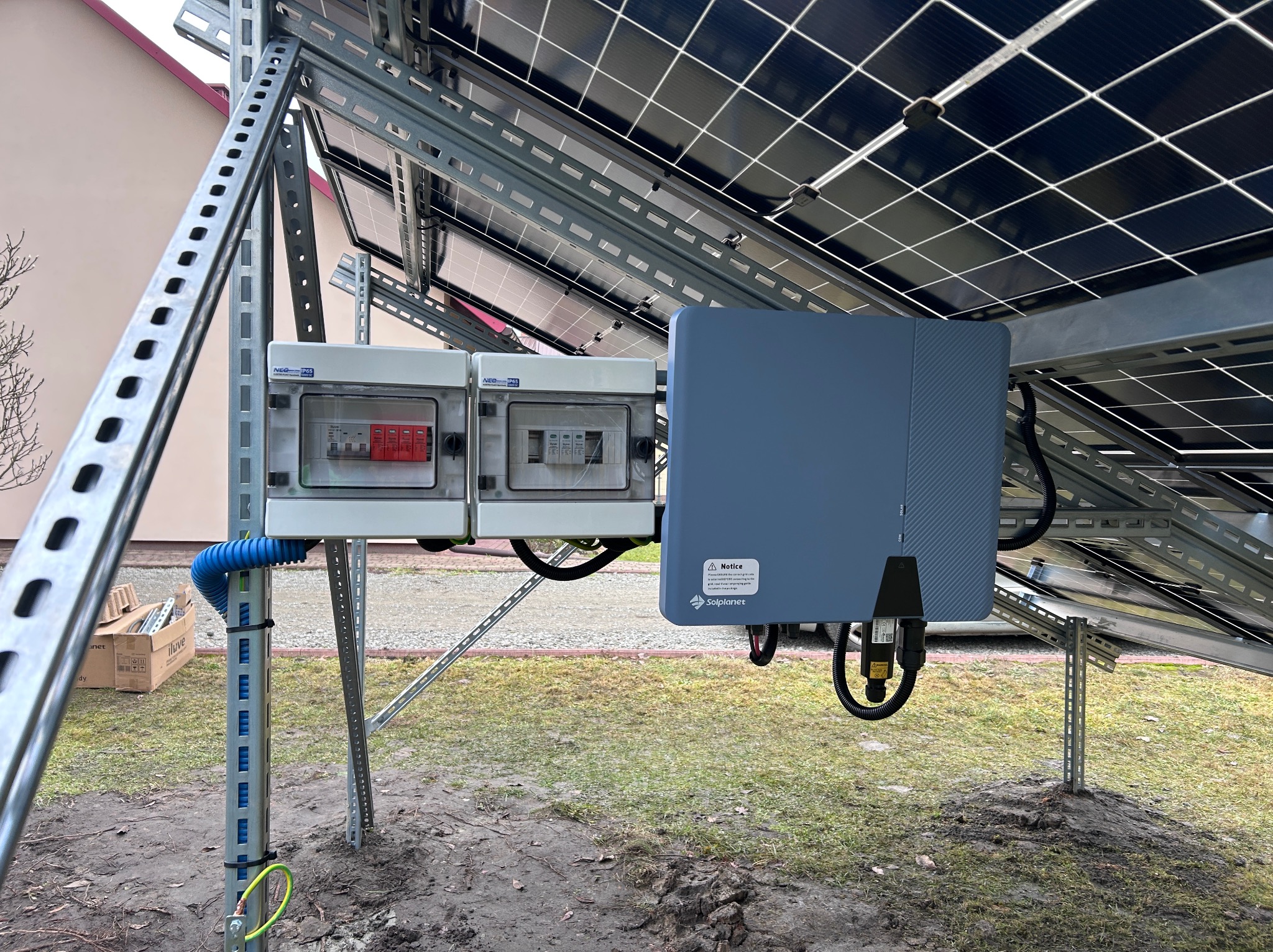 Kompletna instalacja paneli fotowoltaicznych z szarą skrzynką inwertera i dwoma białymi rozdzielnicami na metalowej konstrukcji, widok z dołu.