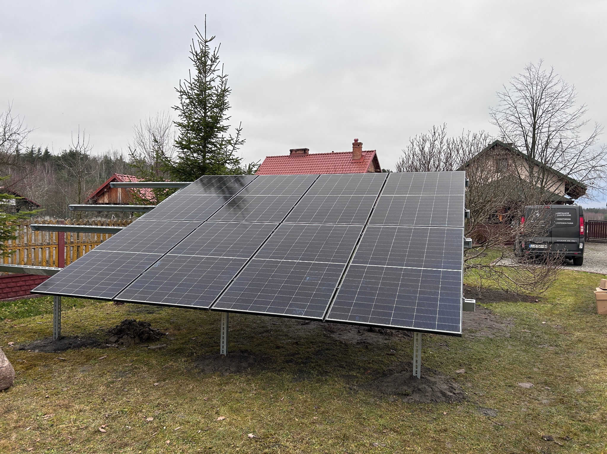 Panele fotowoltaiczne na trawniku w Harasiukach, zamontowane na metalowej konstrukcji. W tle domy i samochód dostawczy z logo firmy.
