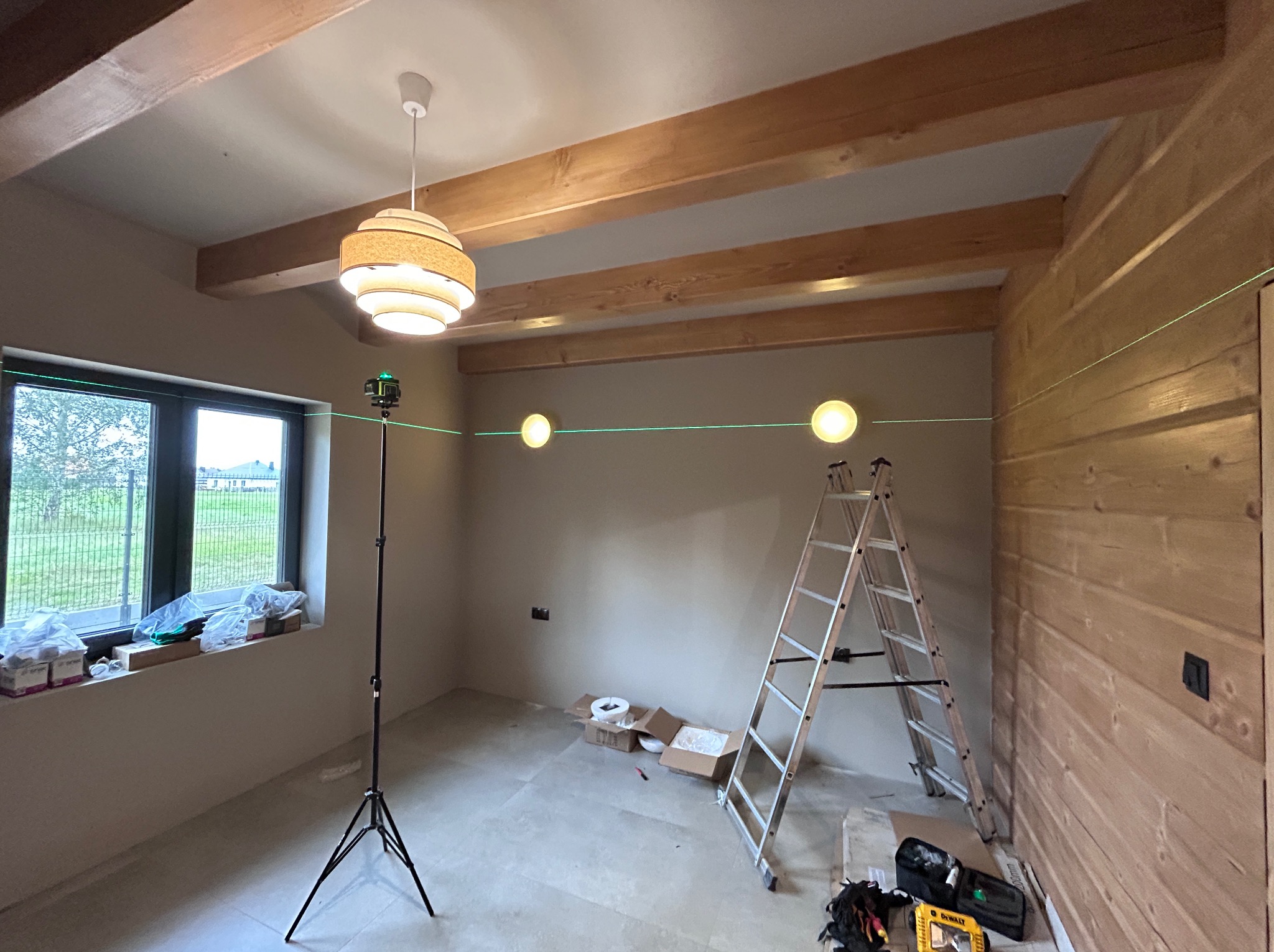 Wnętrze z drewnianymi belkami stropowymi i lampą. Widoczny laser krzyżowy na statywie, wyznaczający poziom, oraz drabina. Ściana wykończona drewnem.