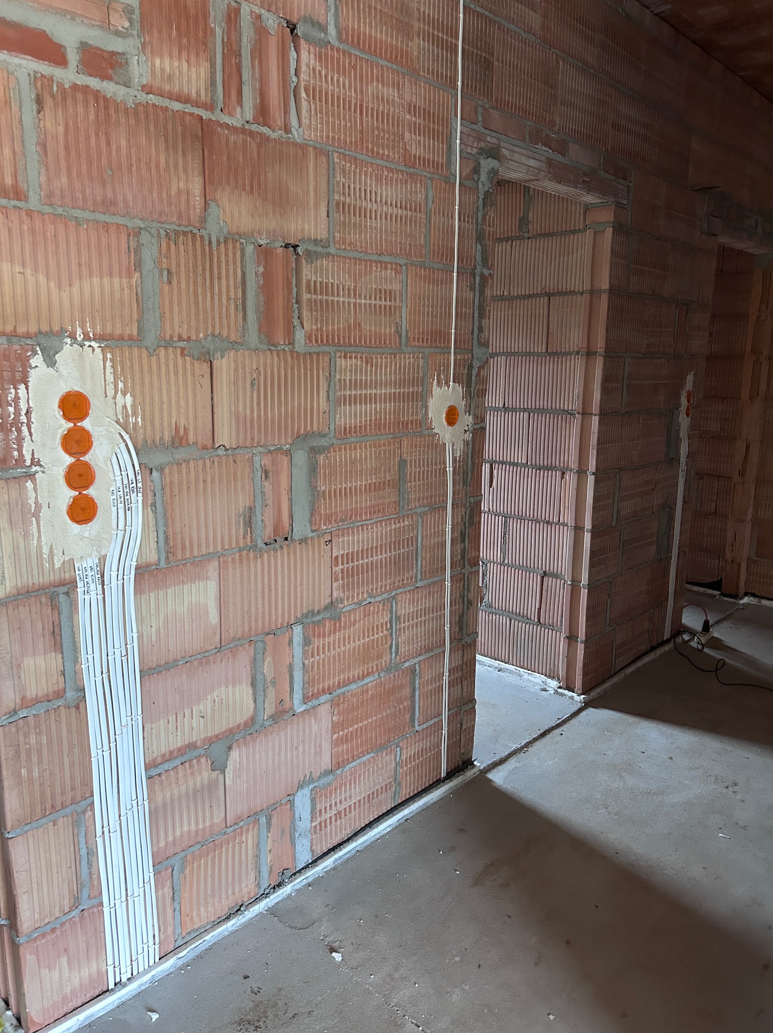 Instalacja elektryczna w trakcie montażu w nowym budynku z czerwonej cegły. Widoczne kable w białych listwach i pomarańczowe puszki pod gniazdka.