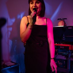 Holiday zespół muzyczny weselny obsługa wesel eventów - Wokalistka w czarnej sukience śpiewa do mikrofonu na scenie z instrumentami klawiszowymi w tle, oświetlona czerwonym i niebieskim światłem.